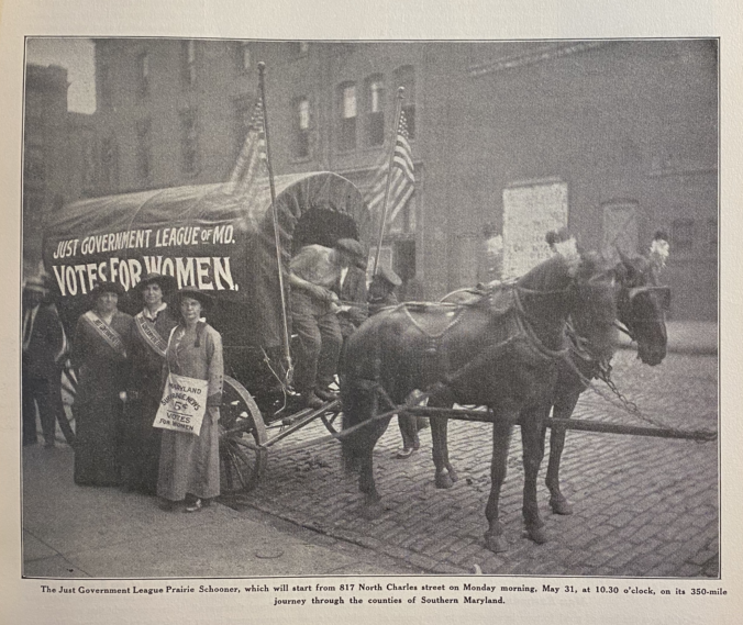 Three women stand at a covered wagon with the words "Just Government League, Votes for Women"