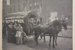 Three women stand at a covered wagon with the words "Just Government League, Votes for Women"