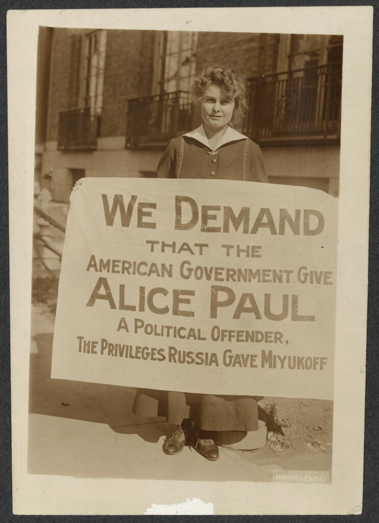 Informal portrait, full-length, Lucy Branham, facing forward, standing in front of a building and holding a sign, 