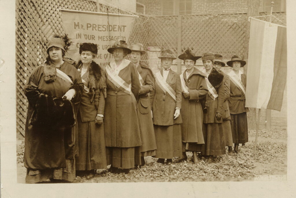 none suffragists lined up against a latticed fence with a banner addressing President Wilson.