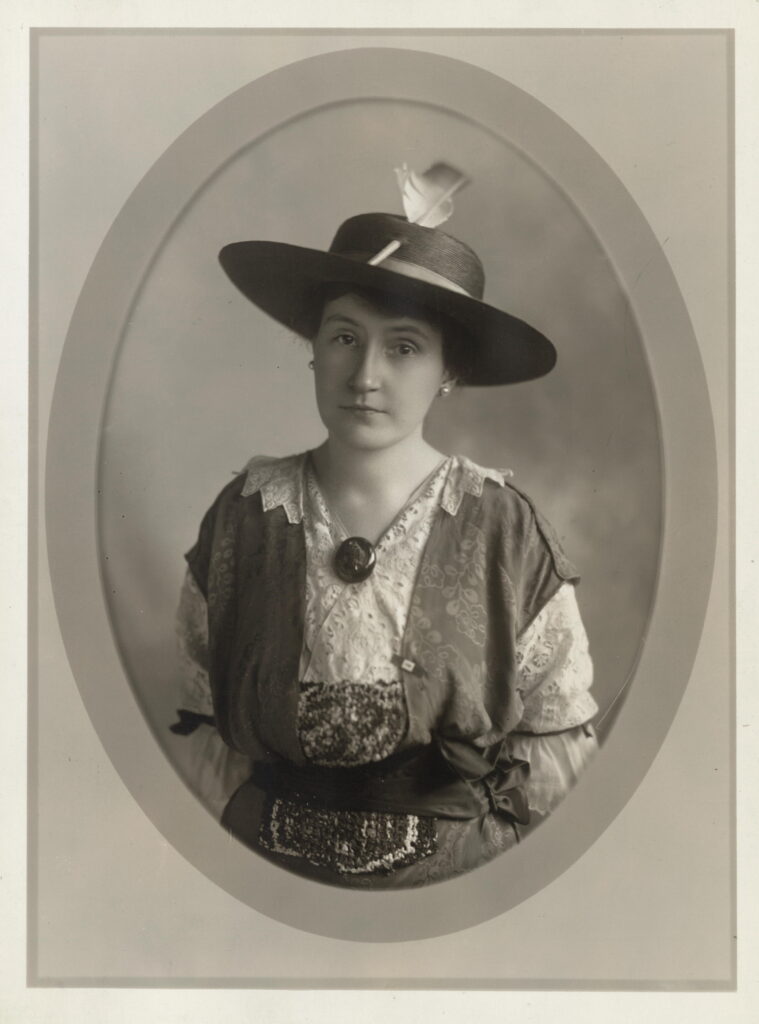 alf length portrait of Edna S. Latimer, facing camera, in hat, with brooch