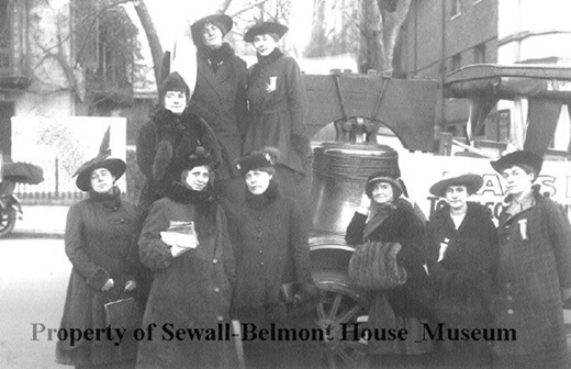 nine women dressed in black coats and hats in front of the Women's Liberty Bell possibly on a float