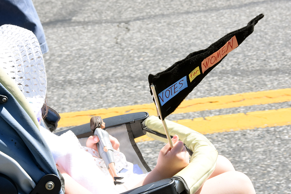 A side shot of a child in a stroller carrying a homemade votes for women flag and a Kamala Harris doll