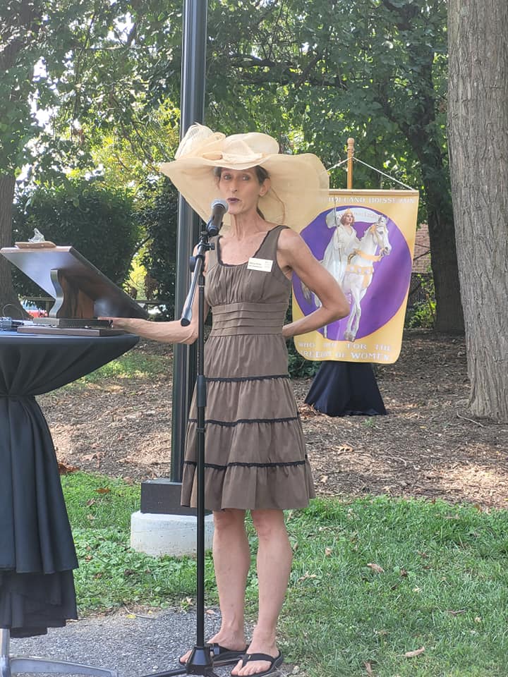 Sandy Spring Museum executive director, Allison Weiss, at a lectern wearing a large straw hat