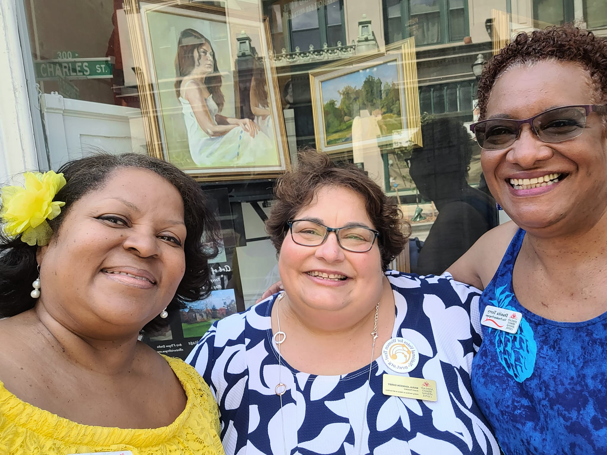 Close up of three women smiling in front of a display window containing artist paintings.