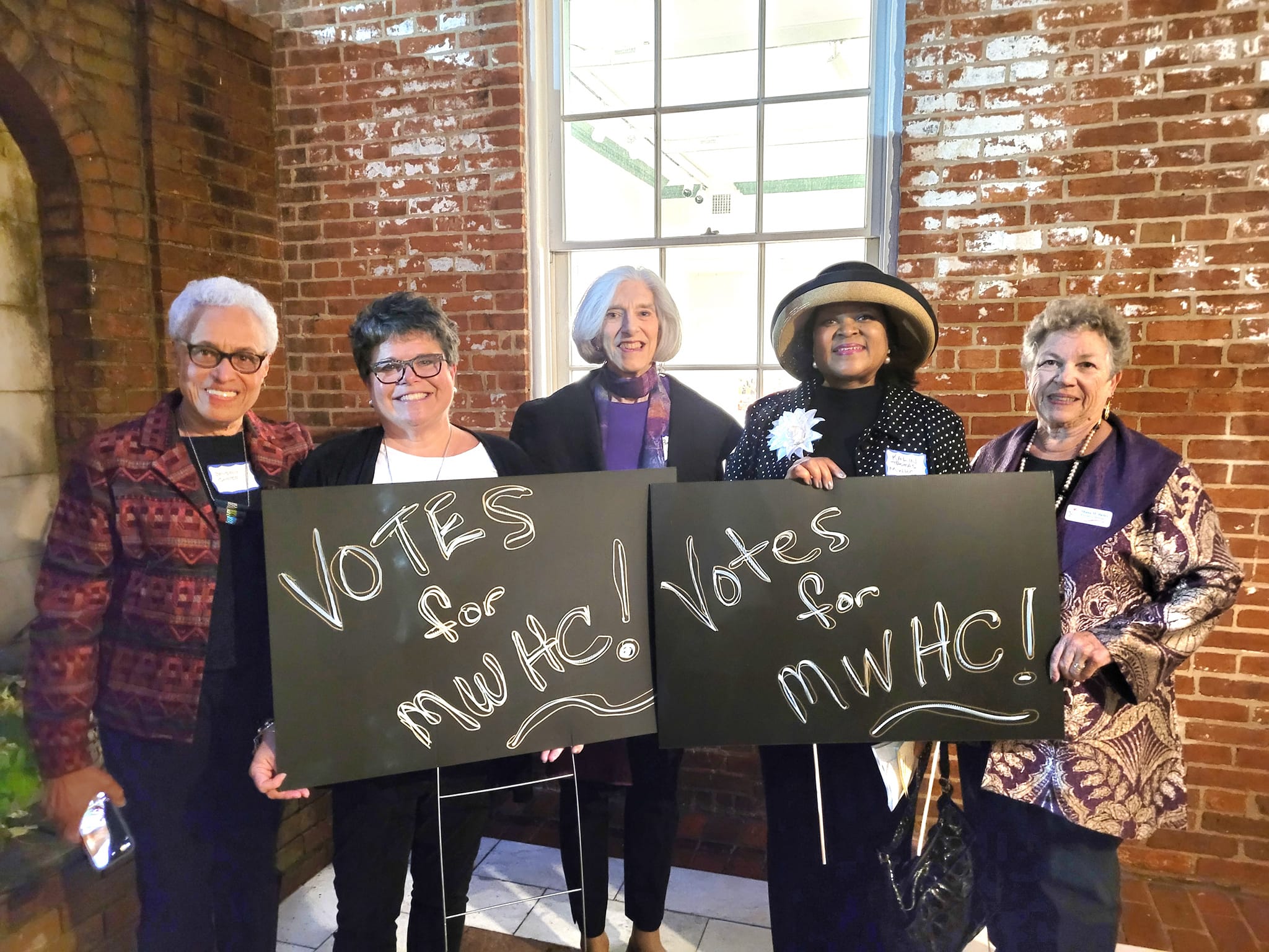 Five MWHC women holding a handmade sign that says "Votes for MWHC"