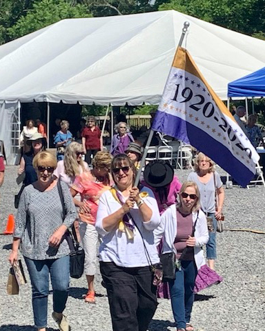 Judy Carbone, carrying a suffrage banner, leading a parade on the day of the Julia Ruhl historical marker unveiling.