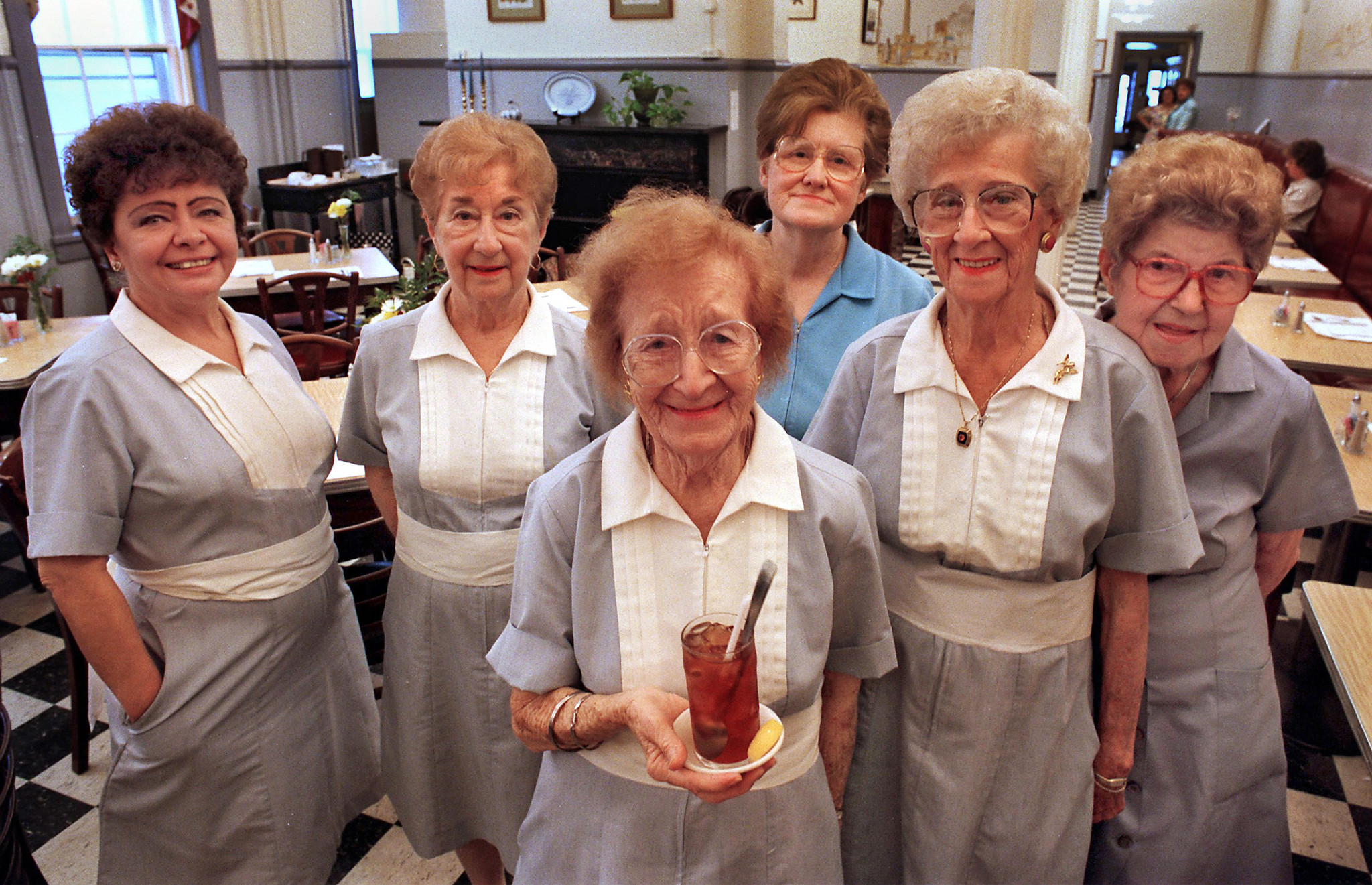 Six WIE waitresses in aline, mid century photo. They are wearing grey uniforms with a white bib-like front