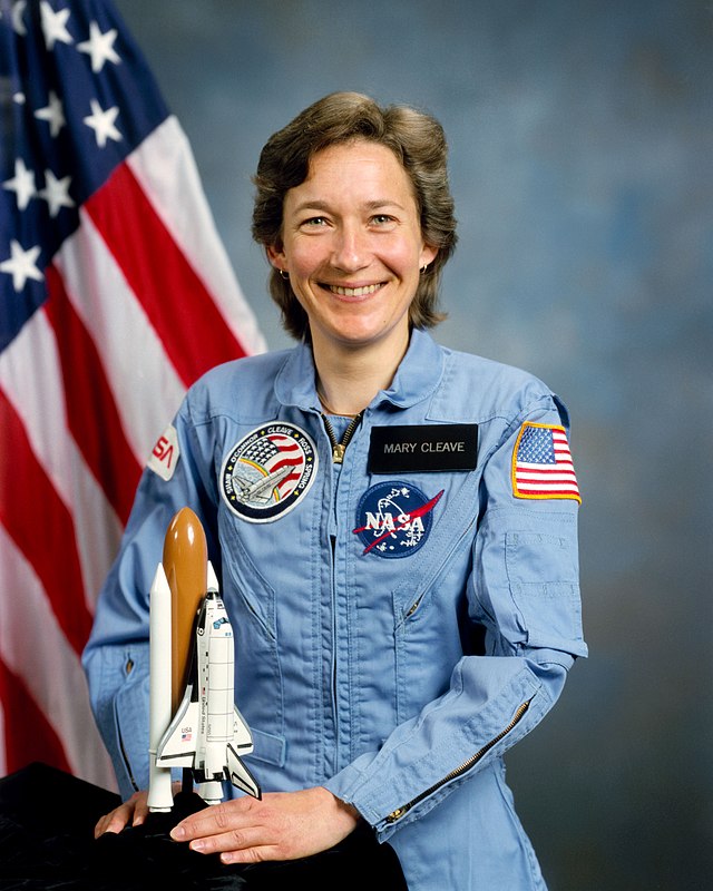 Mary Celave, about 30 years old, posing in a blue flight suit holding a model of the space shuttle
