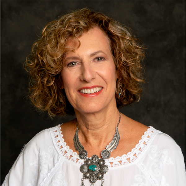 Sandy Cummings headshot against aback background. Wearing a white blouse and large pendent