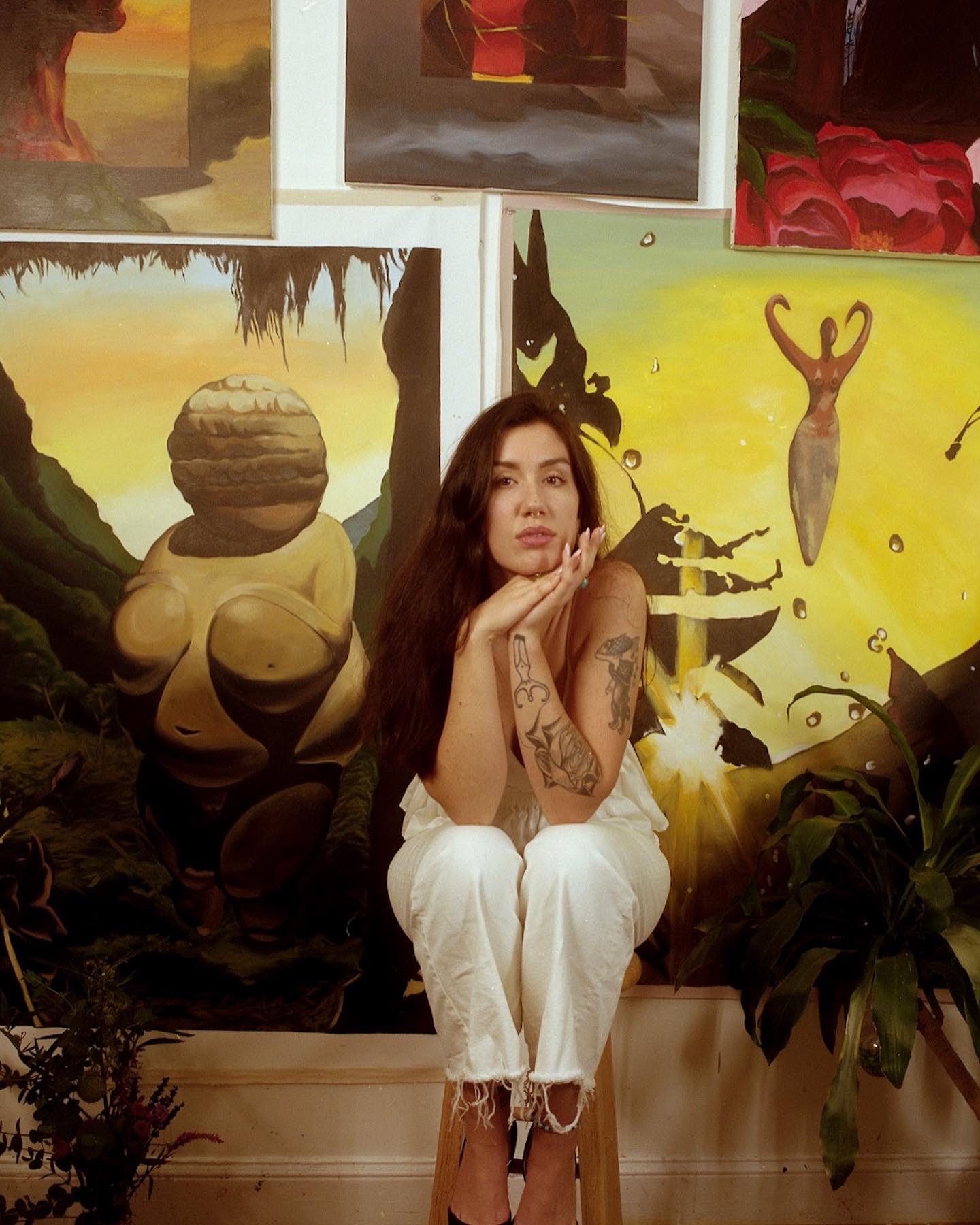 Artist Rowan Bathurst, seated in front of multiple art works. Wearing white jeans and head resting on hands